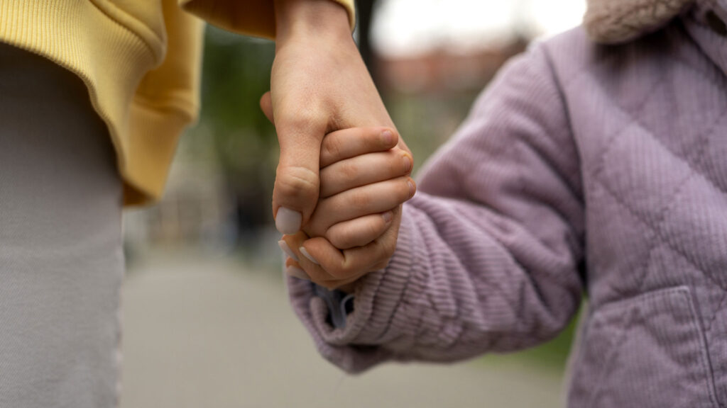 Nahaufnahme: Eine erwachsene Person hält draußen die Hand eines Kindes; beide tragen Jacken, der Hintergrund ist unscharf.
